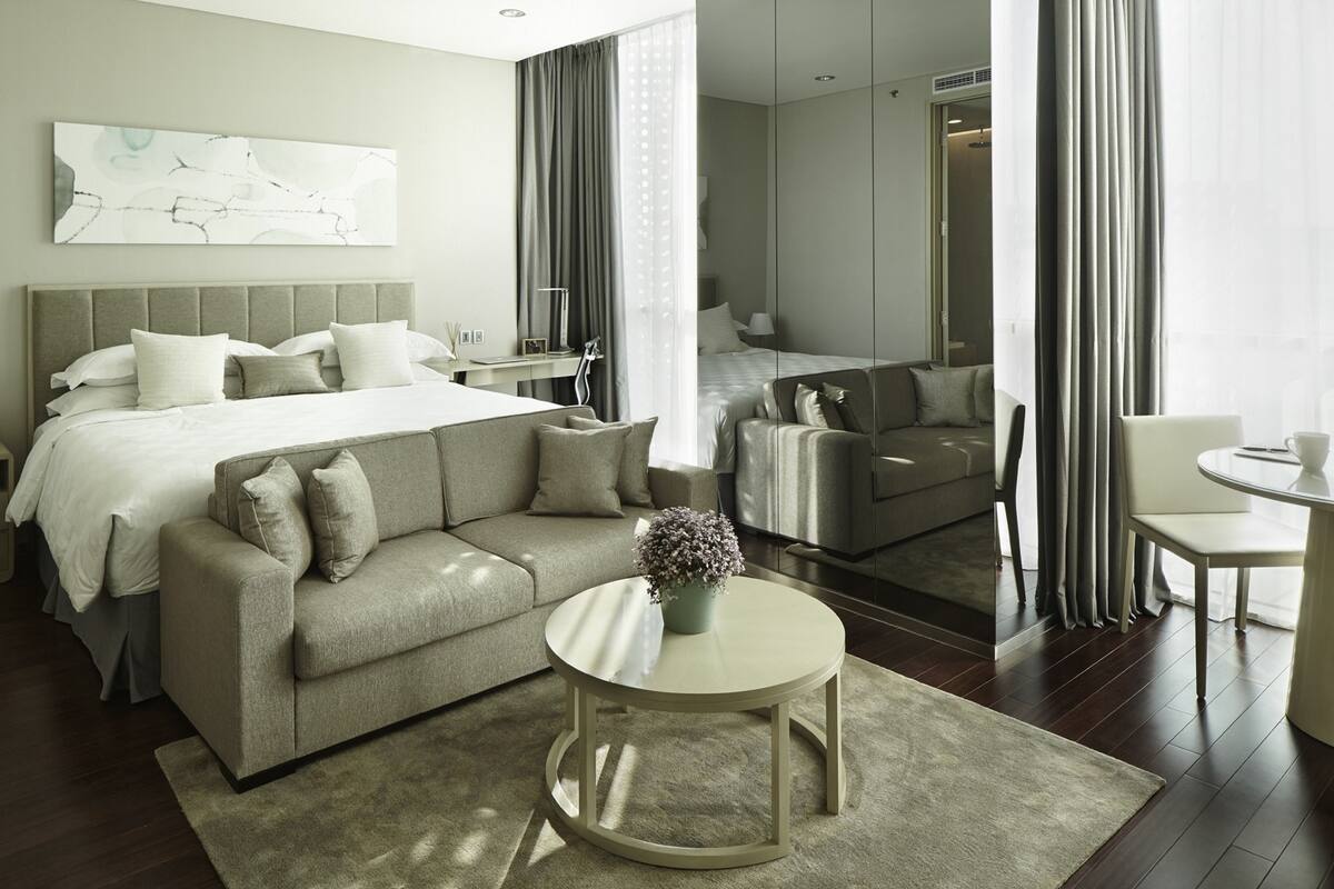 Quarto de hotel moderno com uma cama grande, um sofá, uma mesinha redonda e vista para o quarto vizinho.