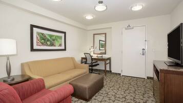 Quarto de hotel com sofá bege, poltrona vermelha, mesa pequena com abajur, escrivaninha com cadeira, televisão e tapete estampado.