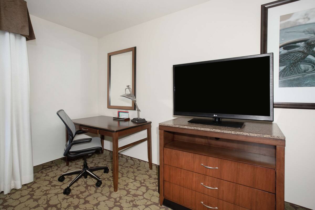 Habitación de hotel con un escritorio de madera, una silla, un televisor sobre un mueble de madera, un espejo y un cuadro enmarcado en la pared.