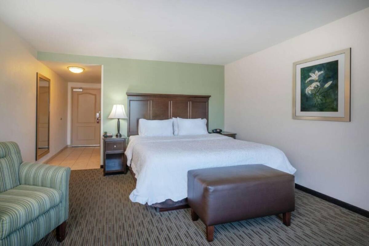 A hotel room with a bed, a chair, a nightstand, a lamp, and a painting on the wall.