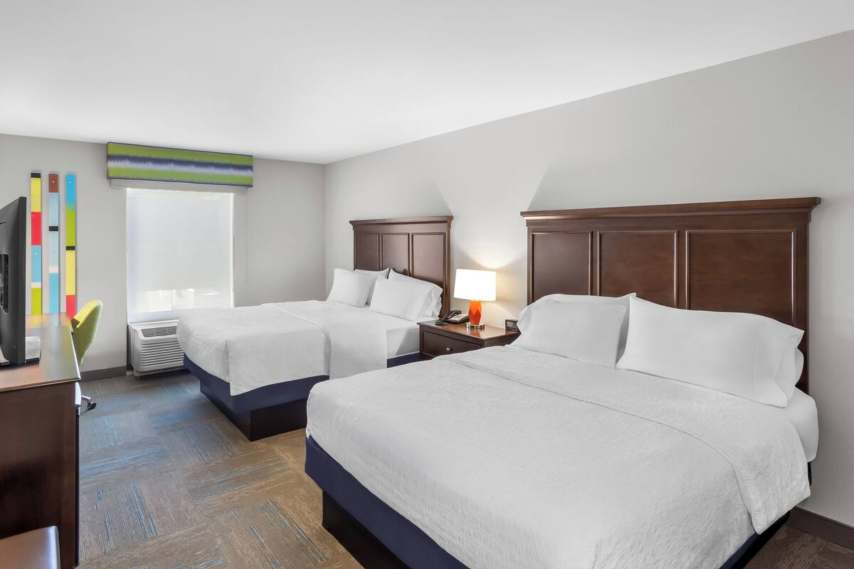 A hotel room with two beds, a desk with a computer, a TV, and a window with blinds.