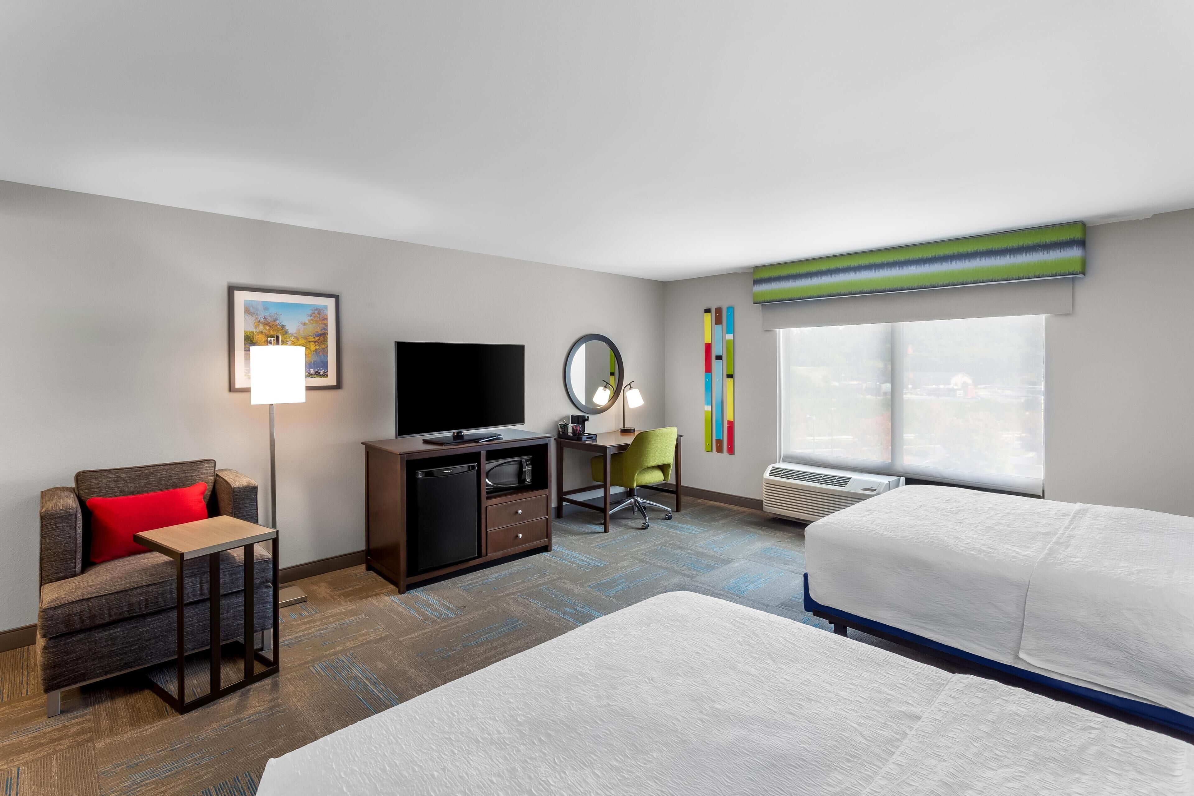 A hotel room with a bed, a desk, a television, a chair, a small table, a mirror, and a window with blinds.