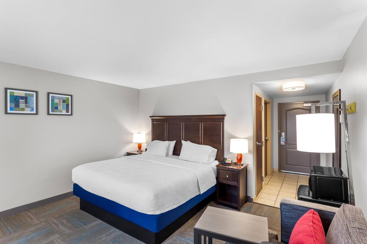 A hotel room with a large bed, bedside tables with lamps, a small table, a chair, and a door leading to a bathroom.