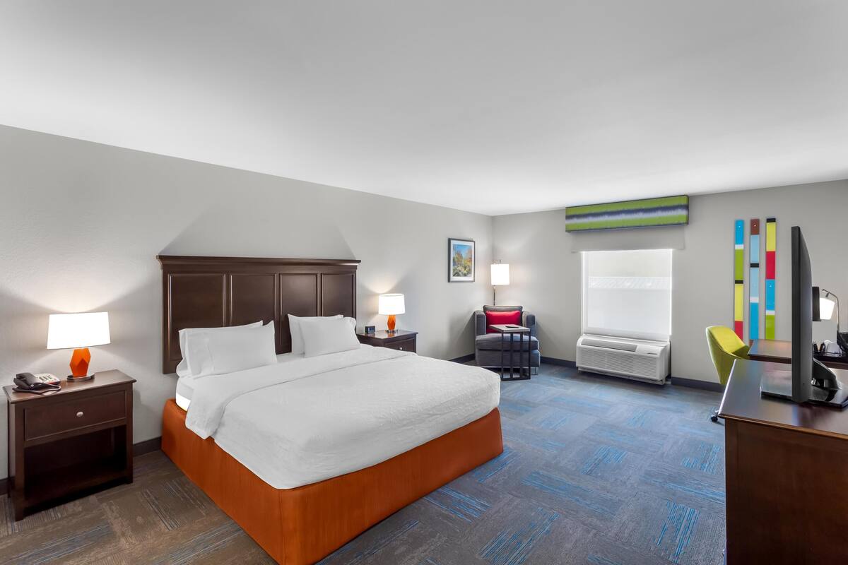 A hotel room with a large bed, bedside tables with lamps, a desk with a chair, a window, and a wall with colorful decor.