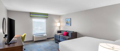 A hotel room with a bed, a desk with a TV, a chair, a window with a view, and a lamp.