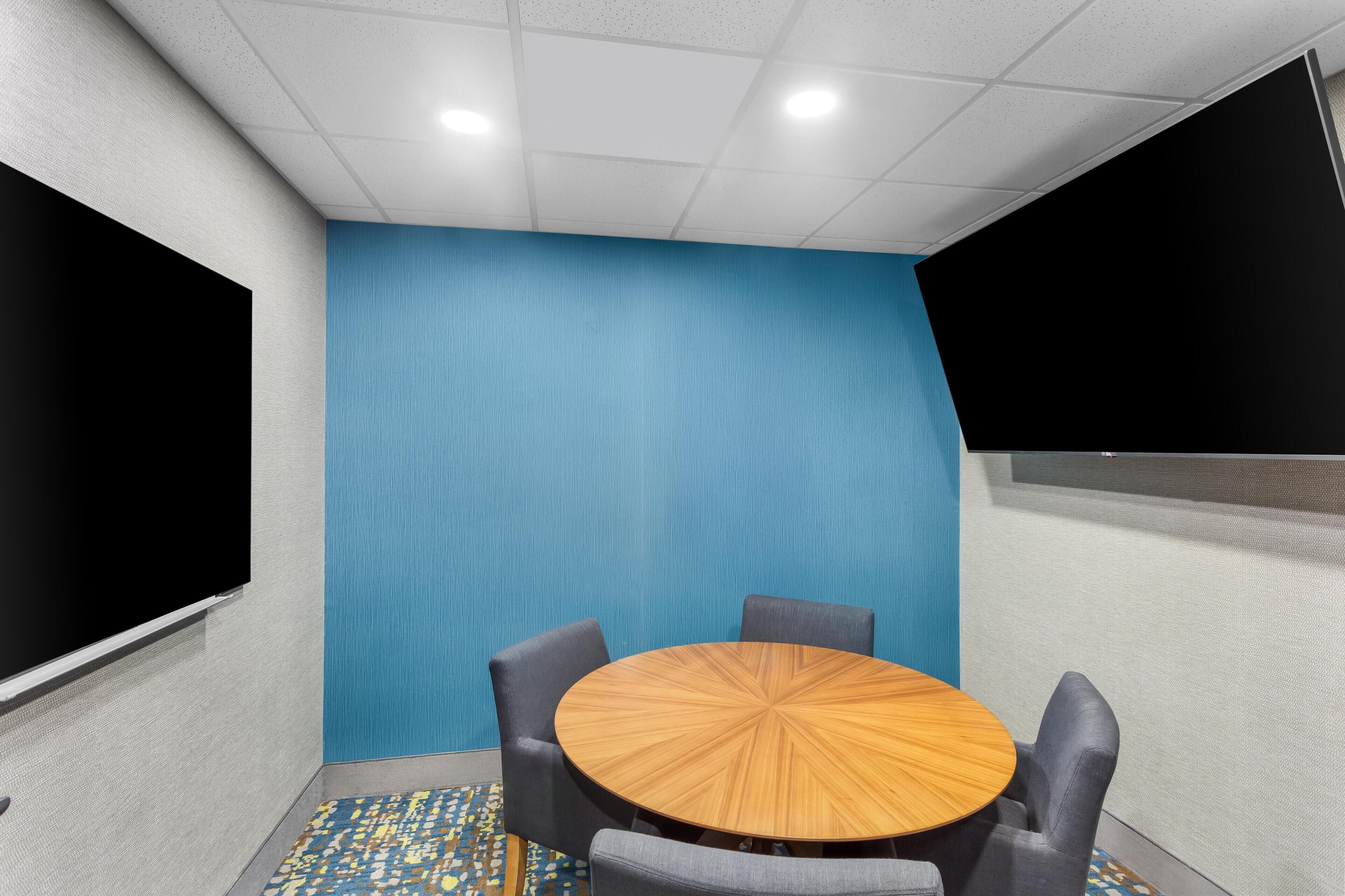 A conference room with a round wooden table, four grey chairs, a blue textured wall, and a large black screen on one side.