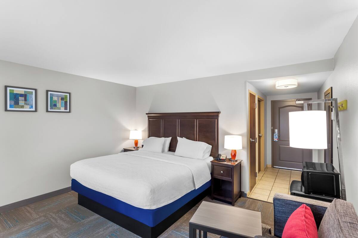 A hotel room with a large bed, two bedside tables with lamps, a small table, a chair, and a door leading to a bathroom.