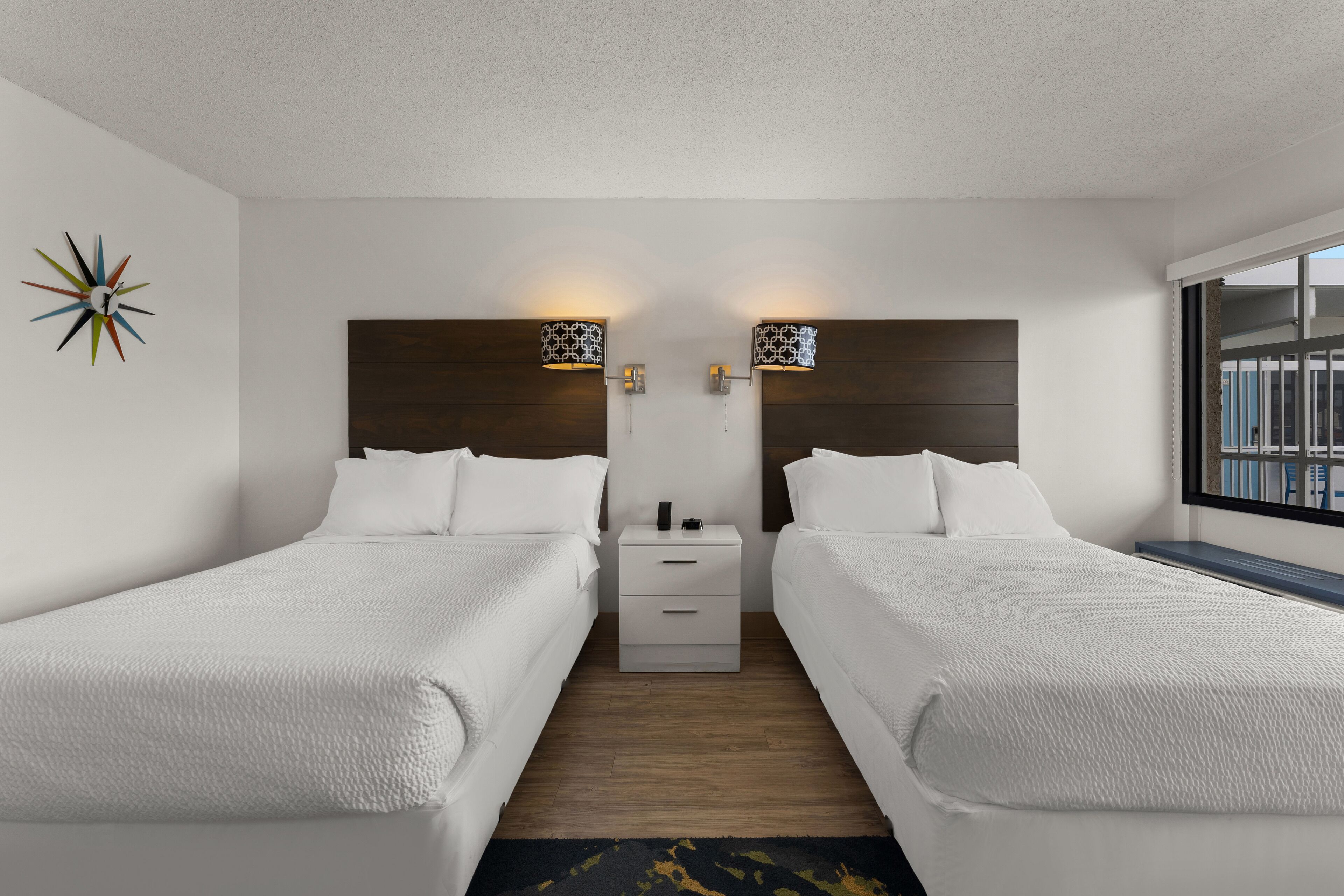 A hotel room with two beds, a nightstand, and a wall-mounted lamp.