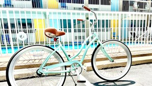 Un vélo turquoise est garé sur une surface en béton, délimitée par une ligne blanche, devant une clôture blanche. On aperçoit une piscine et des bâtiments colorés en arrière-plan.