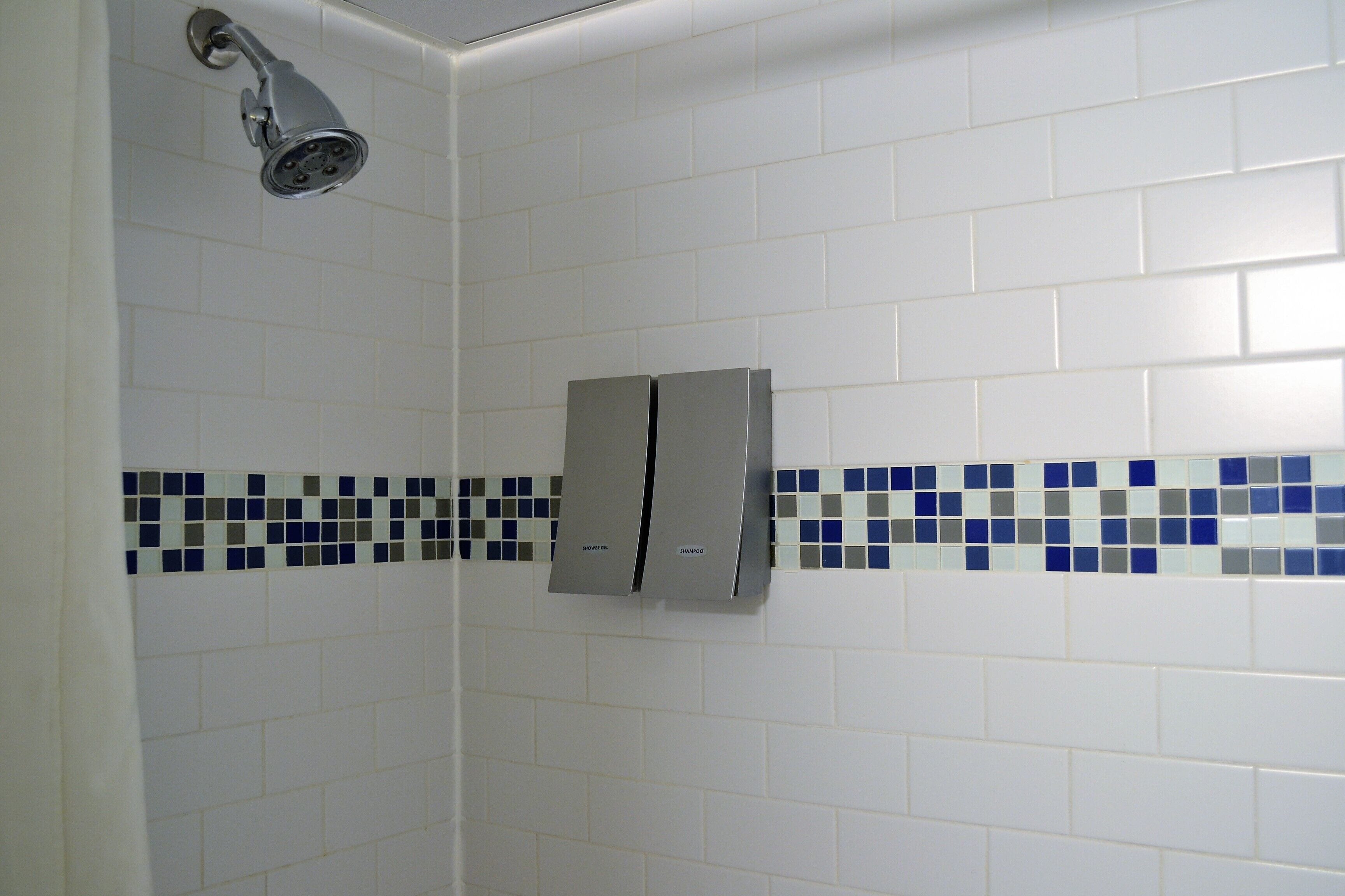A bathroom with white tiles, a showerhead, and a soap dispenser.