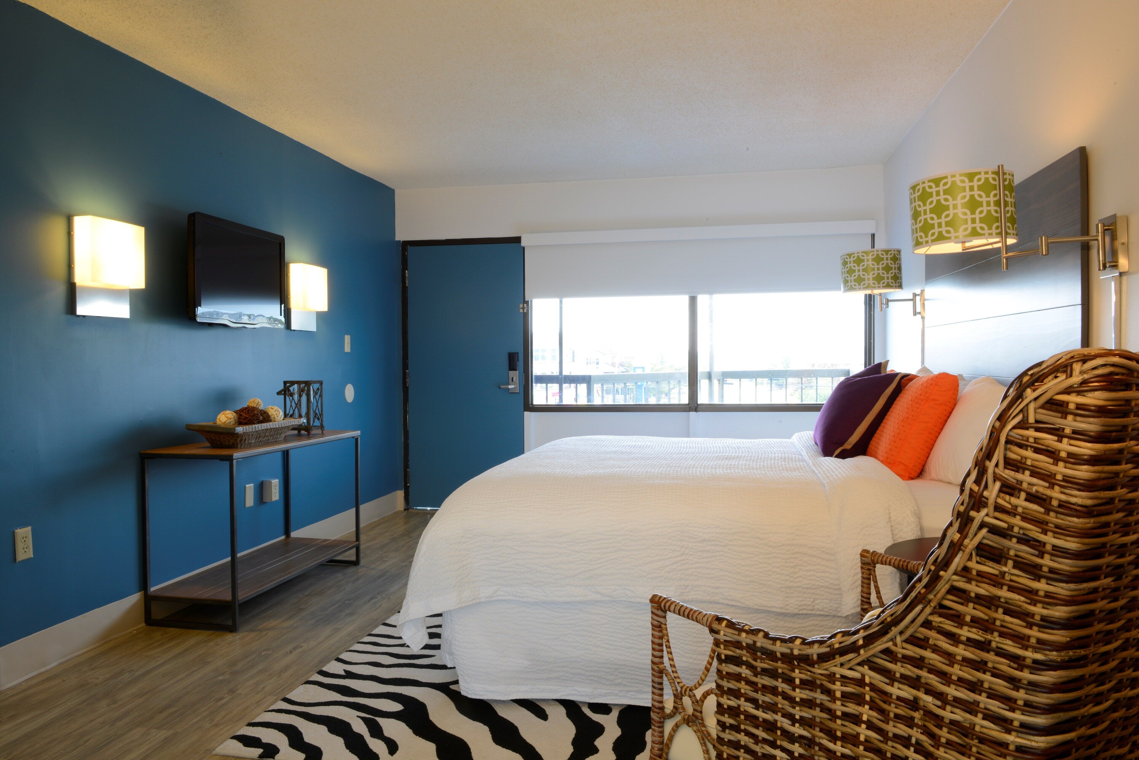 A bedroom with a large bed, a wicker chair, a console table with a basket, and two wall-mounted lamps.