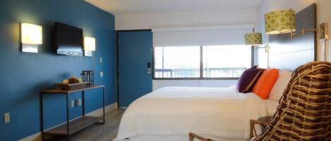 A bedroom with a large bed, a wicker chair, a console table with a basket, and two wall-mounted lamps.