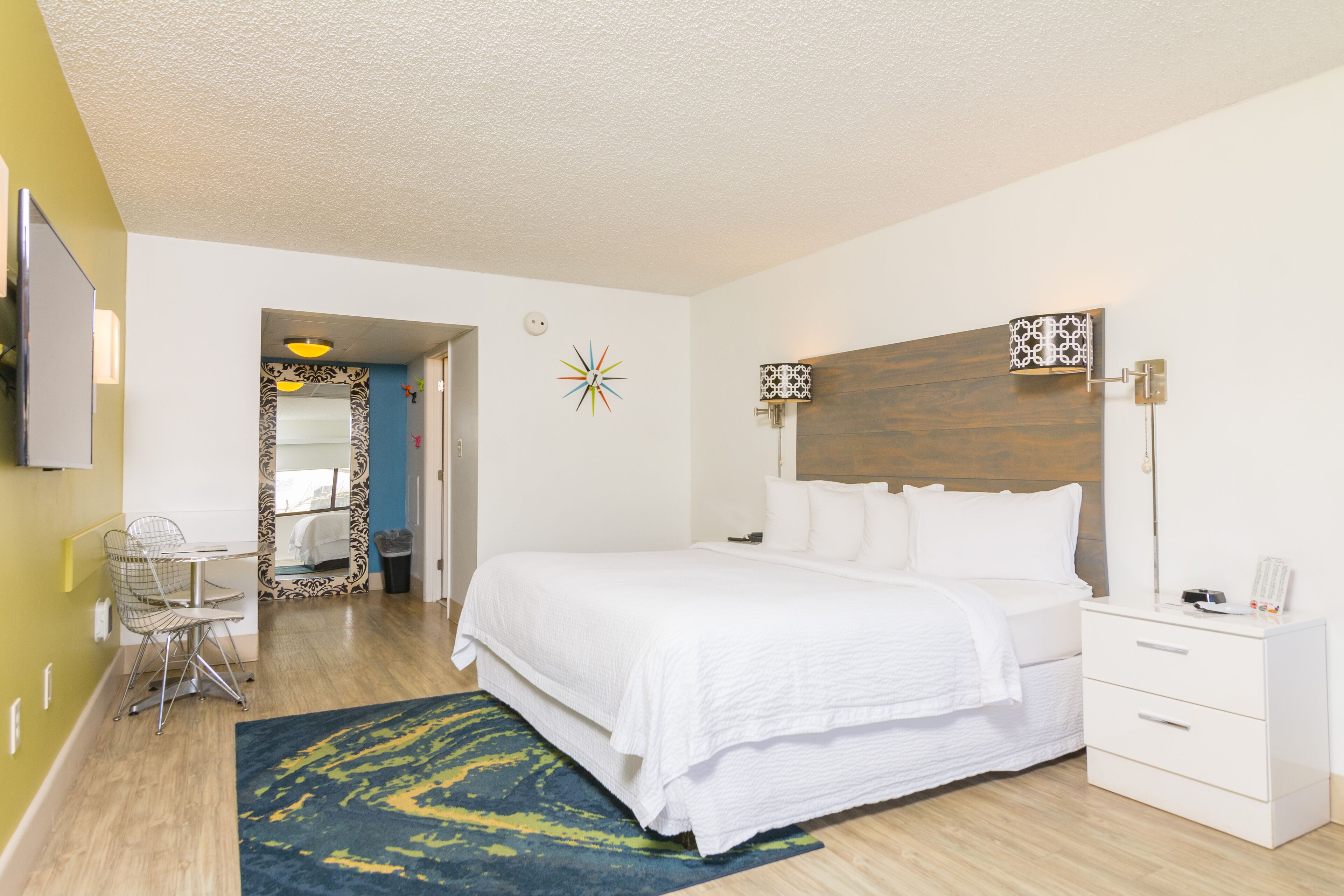 Un dormitorio con una cama, mesita de noche y una silla. Hay un espejo en la pared, un reloj y una puerta que da a otra habitación.