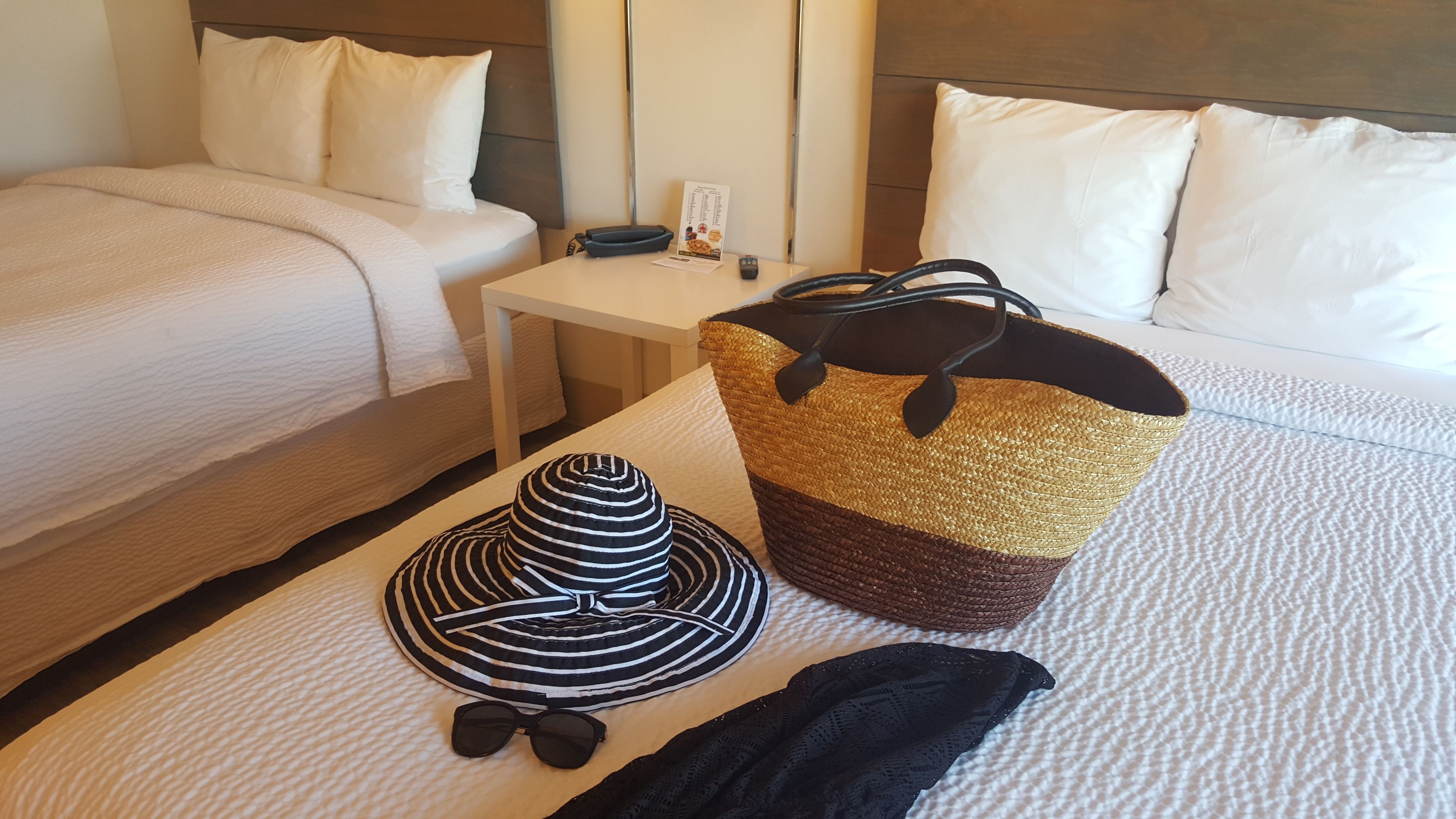 Una cama bien hecha con sábanas blancas, una mesita de noche con un control remoto, un sombrero a rayas, gafas de sol y un bolso tejido.