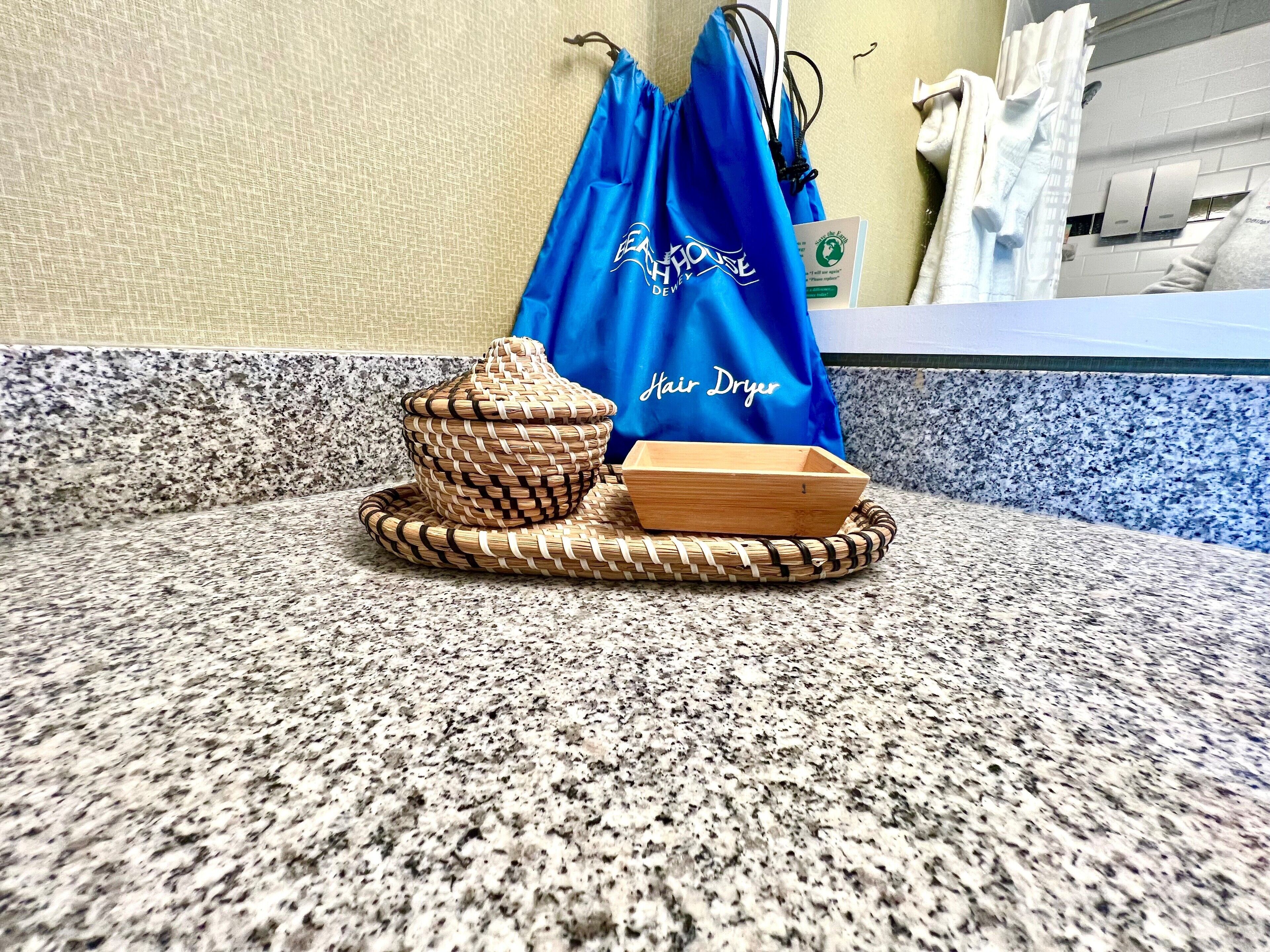 A bathroom counter with a woven basket and a wooden bowl, a blue bag with "Hair Dryer" text, and a towel rack in the background.