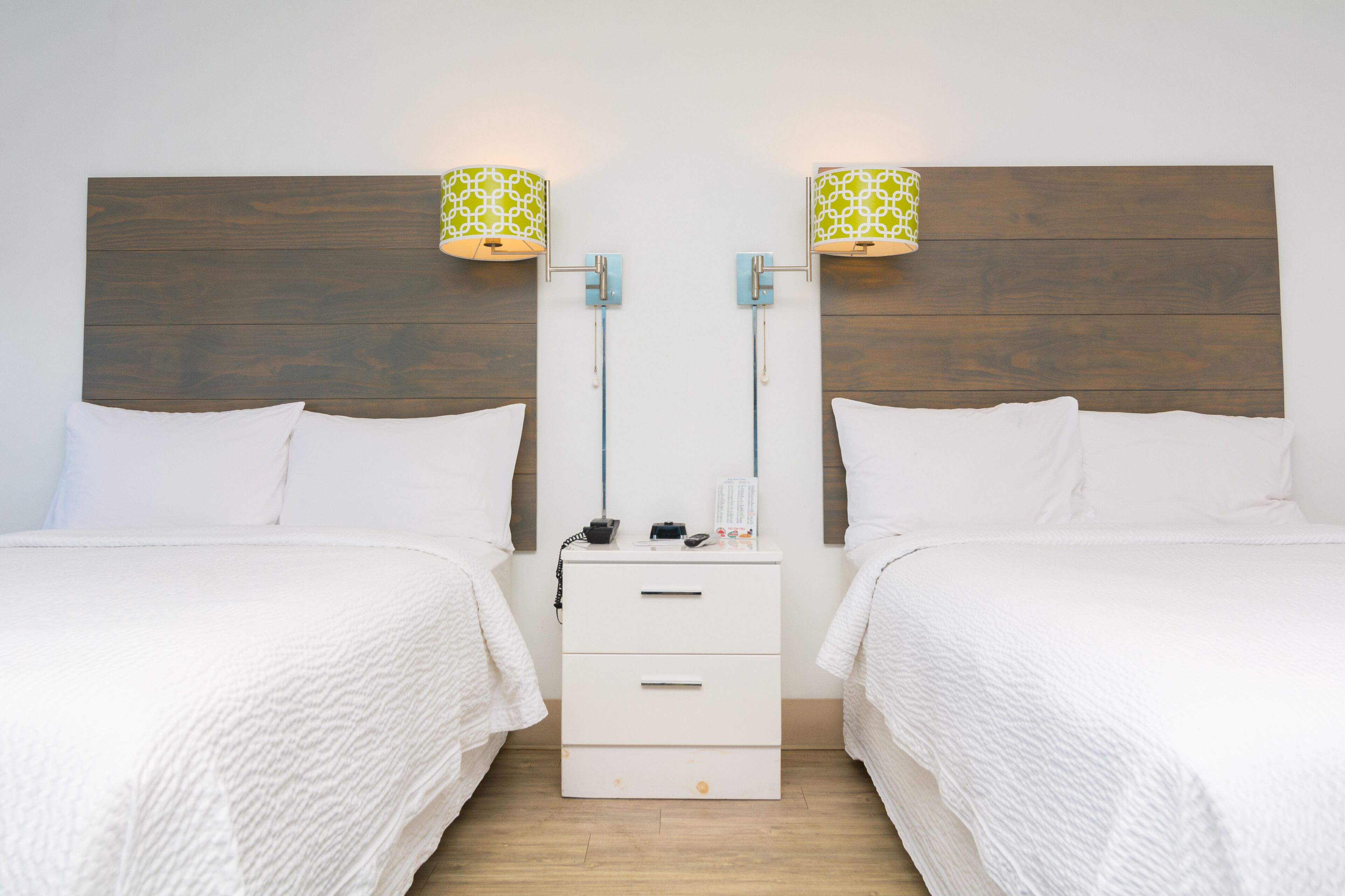 Une chambre d’hôtel avec deux lits, une tête de lit en bois, une table de chevet avec une lampe et un petit tiroir.