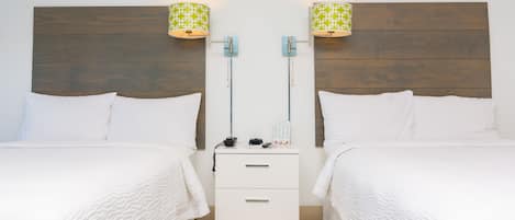 A hotel room with two beds, a wooden headboard, a nightstand with a lamp, and a small drawer.