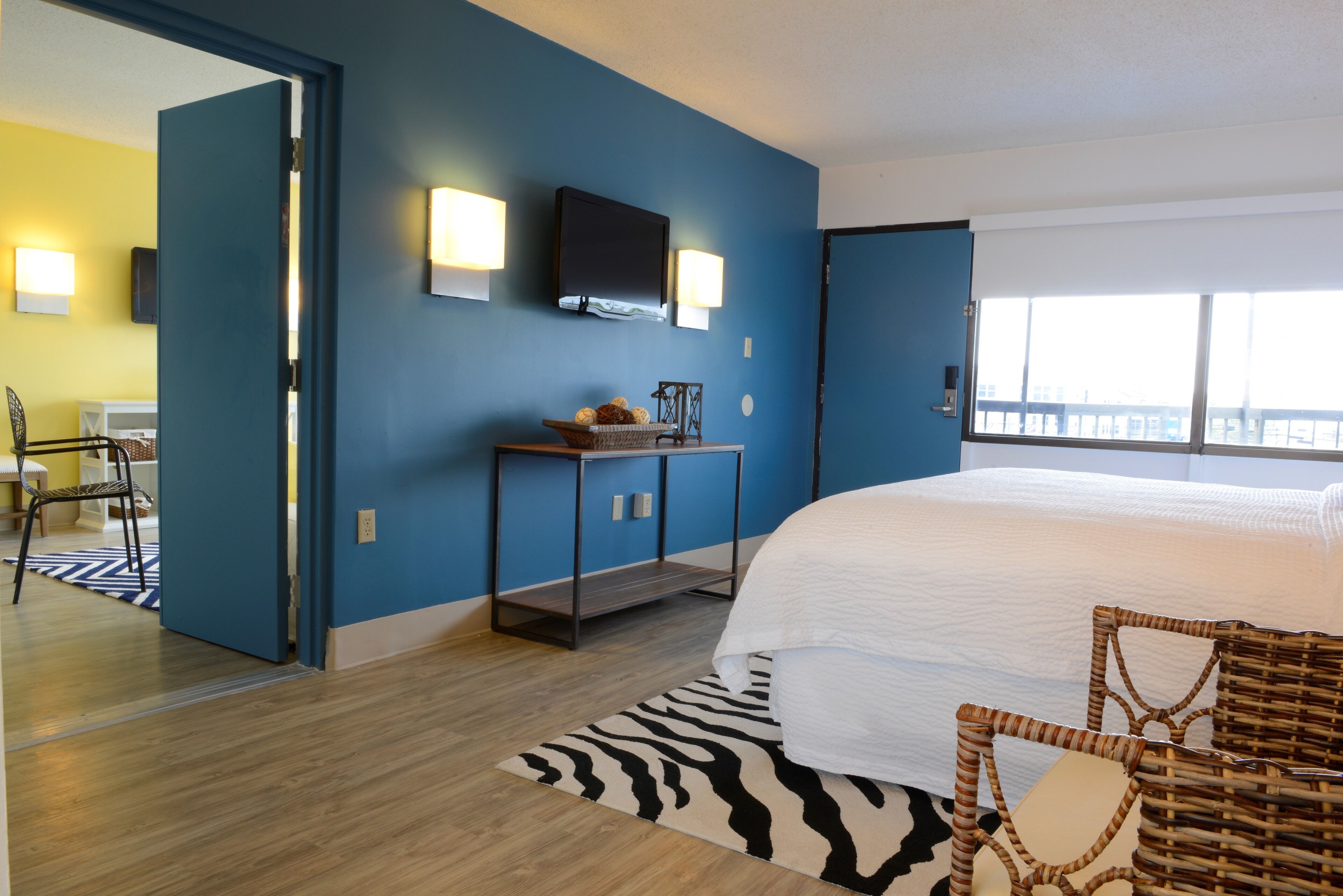 A modern hotel room with a large bed, a flat-screen TV mounted on the wall, a small table with a basket, and a view of the city through large windows.