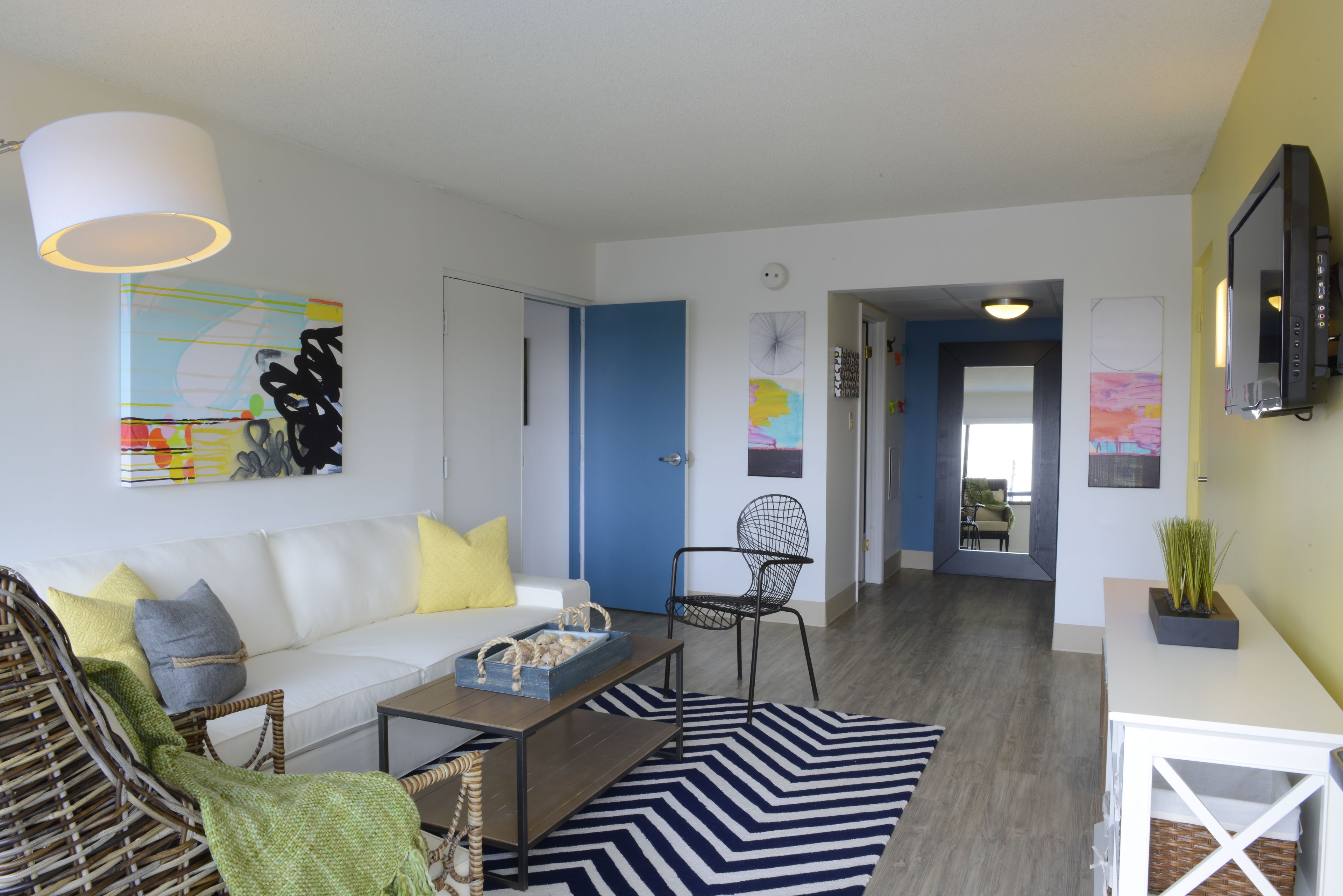Een moderne woonkamer met een witte bank, een rieten stoel, een salontafel en een zwart-wit gestreept tapijt. Er is een blauwe deur die naar een andere kamer leidt, en een televisie hangt aan de muur.