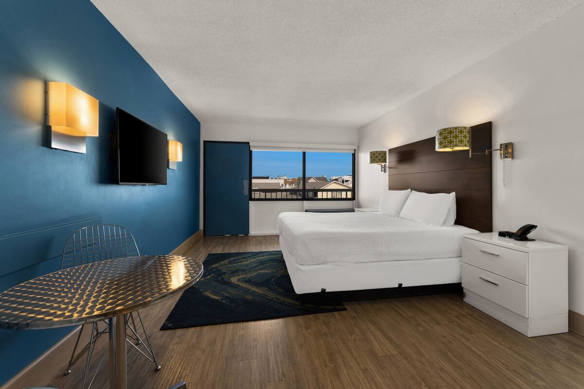 A modern hotel room with a large bed, a desk with a chair, a flat-screen TV, and a view of the city through large windows.