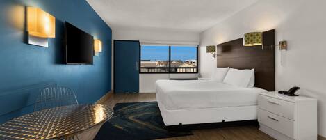 A modern hotel room with a large bed, a desk with a chair, a flat-screen TV, and a view of the city through large windows.