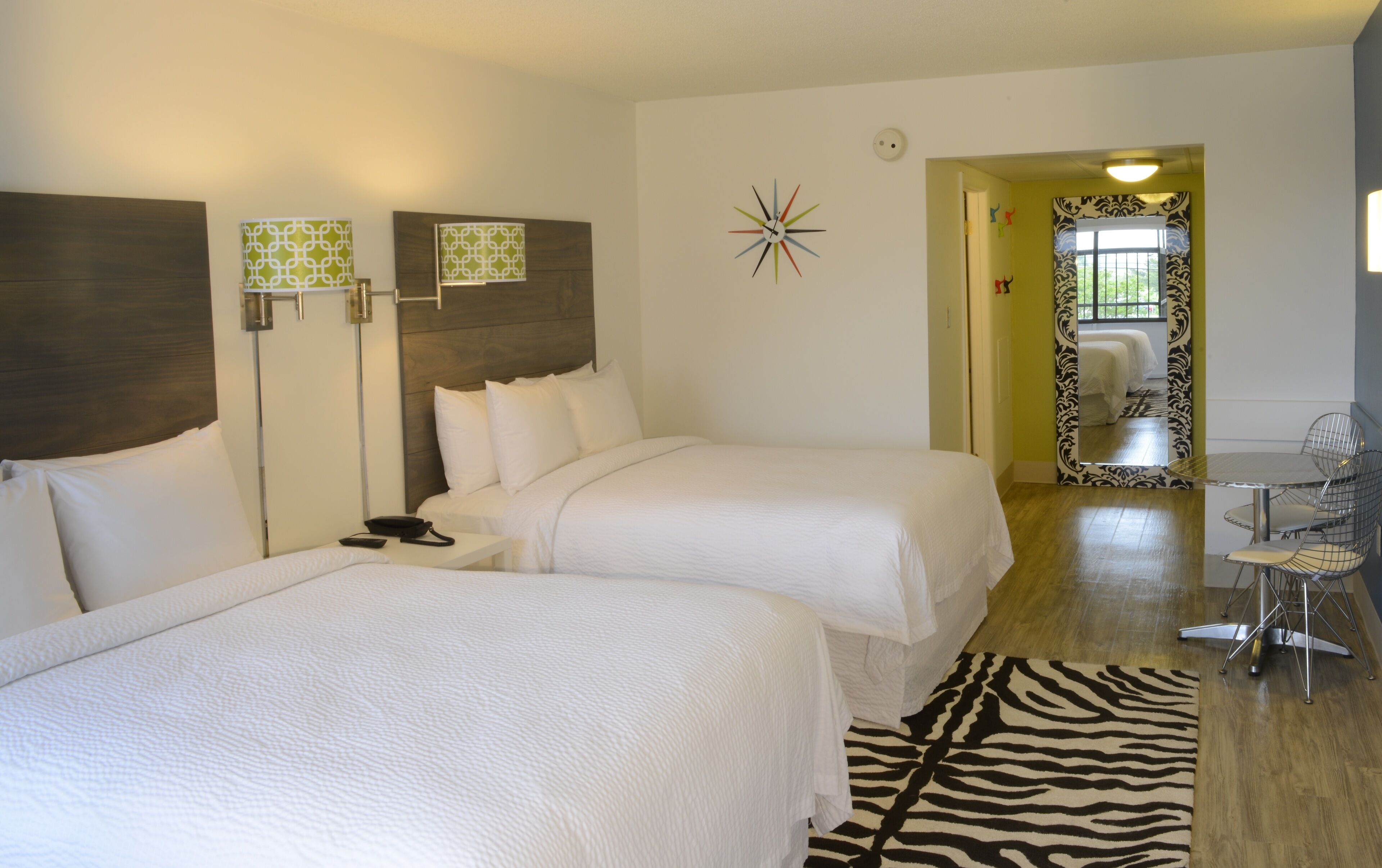 A hotel room with two beds, a zebra-patterned rug, a clock on the wall, and a small table with chairs.