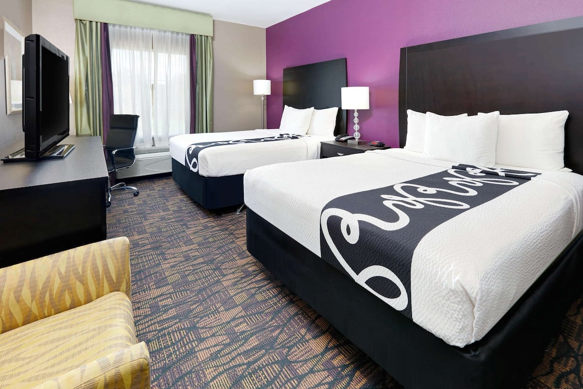 A hotel room with two beds, a desk with a TV, a chair, and a large window with curtains.