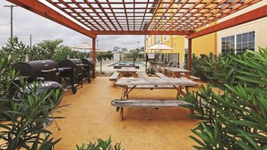 Een terras buiten met houten tafels en banken, een pergola en barbecues.