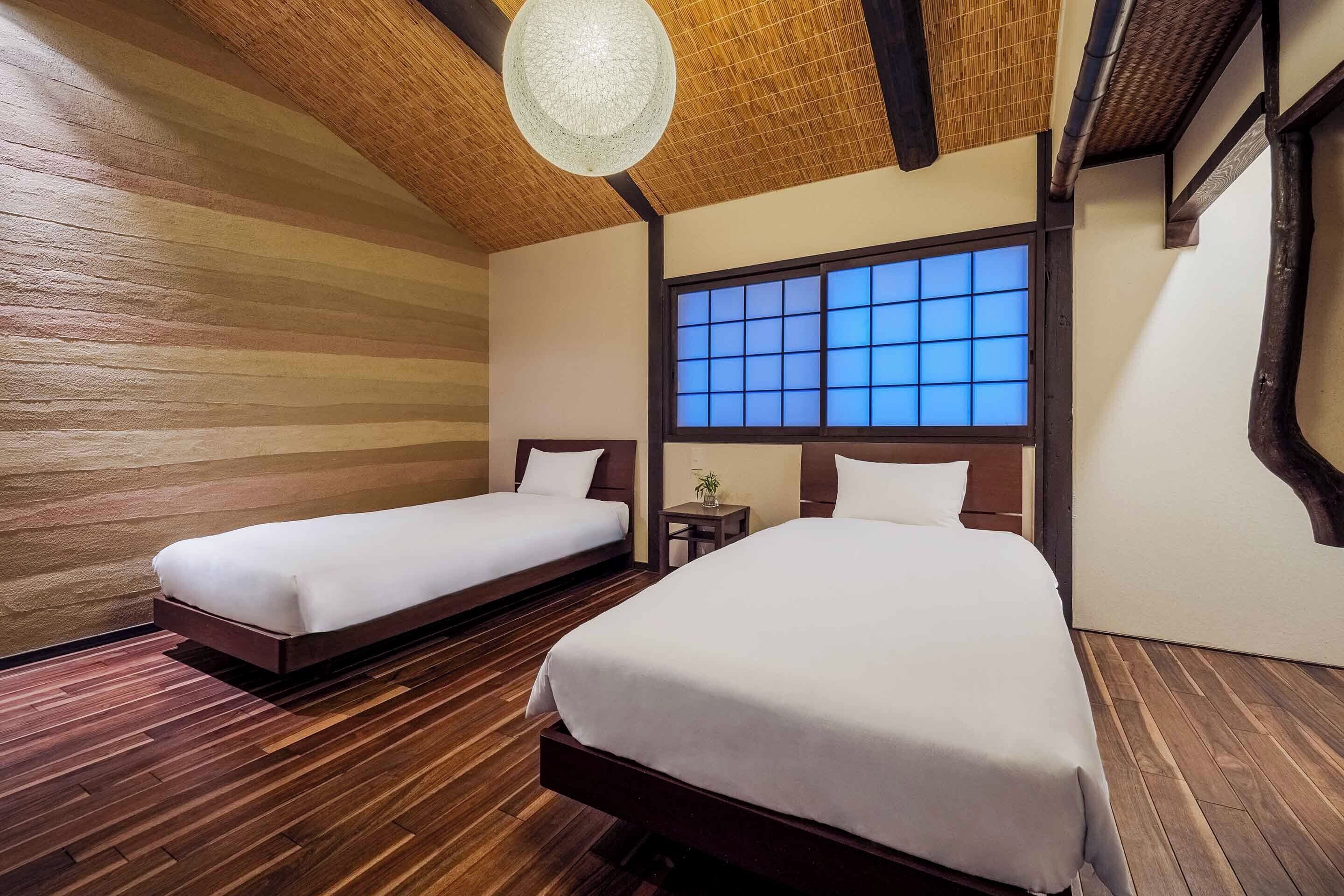 Ein Hotelzimmer mit zwei Betten, Holzboden, einem großen Fenster mit blauer Glasbeschichtung und einer Hängelampe.