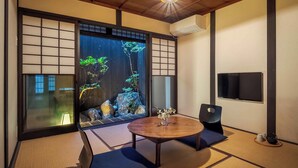 Een traditionele Japanse kamer met tatami vloerbedekking, een lage tafel met een vaas, een zwarte stoel en een televisie aan de muur.