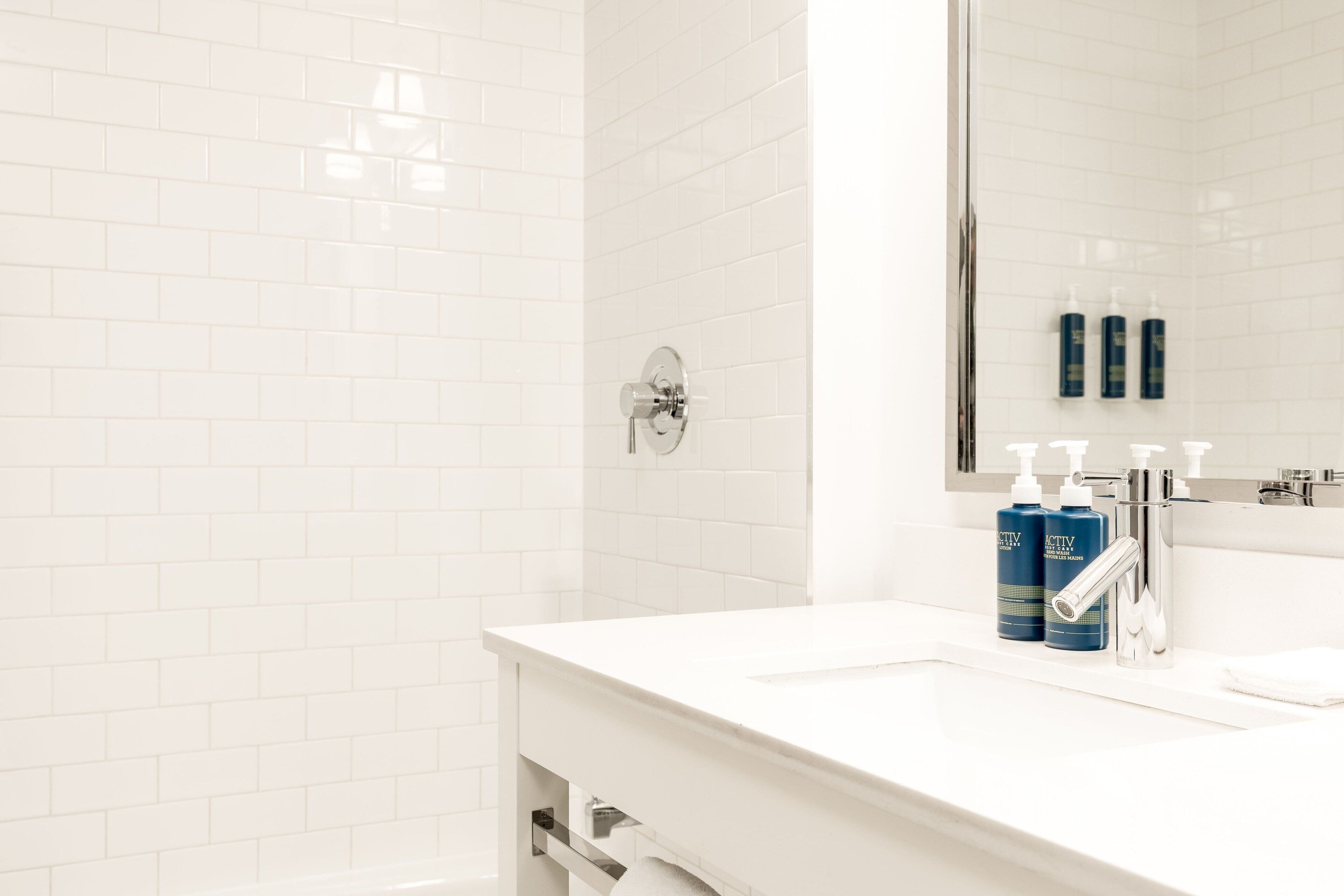 A bathroom with white tiled walls, a white sink, and two blue bottles of lotion on the counter.