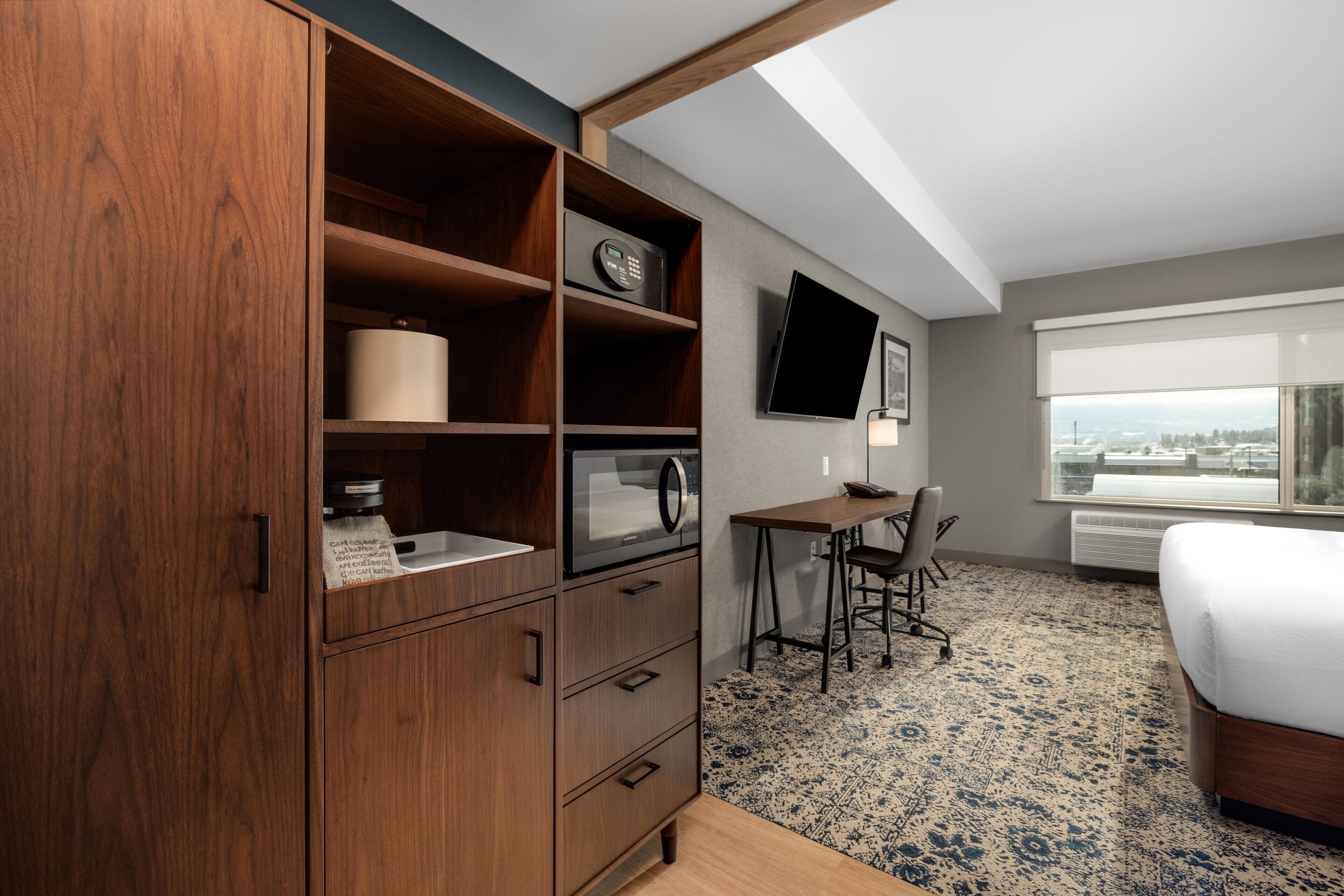 Habitación de hotel con un armario de madera, un escritorio con silla, una cama, un televisor de pantalla plana montado en la pared y un ventanal con vistas a la ciudad.