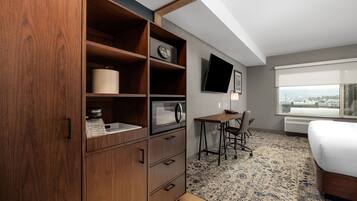 Habitación de hotel con un armario de madera, un escritorio con silla, una cama, un televisor de pantalla plana montado en la pared y un ventanal con vistas a la ciudad.