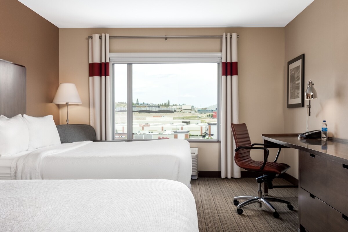 Habitación de hotel con una cama grande, un escritorio, una silla, una lámpara y vistas a la ciudad a través de la ventana.
