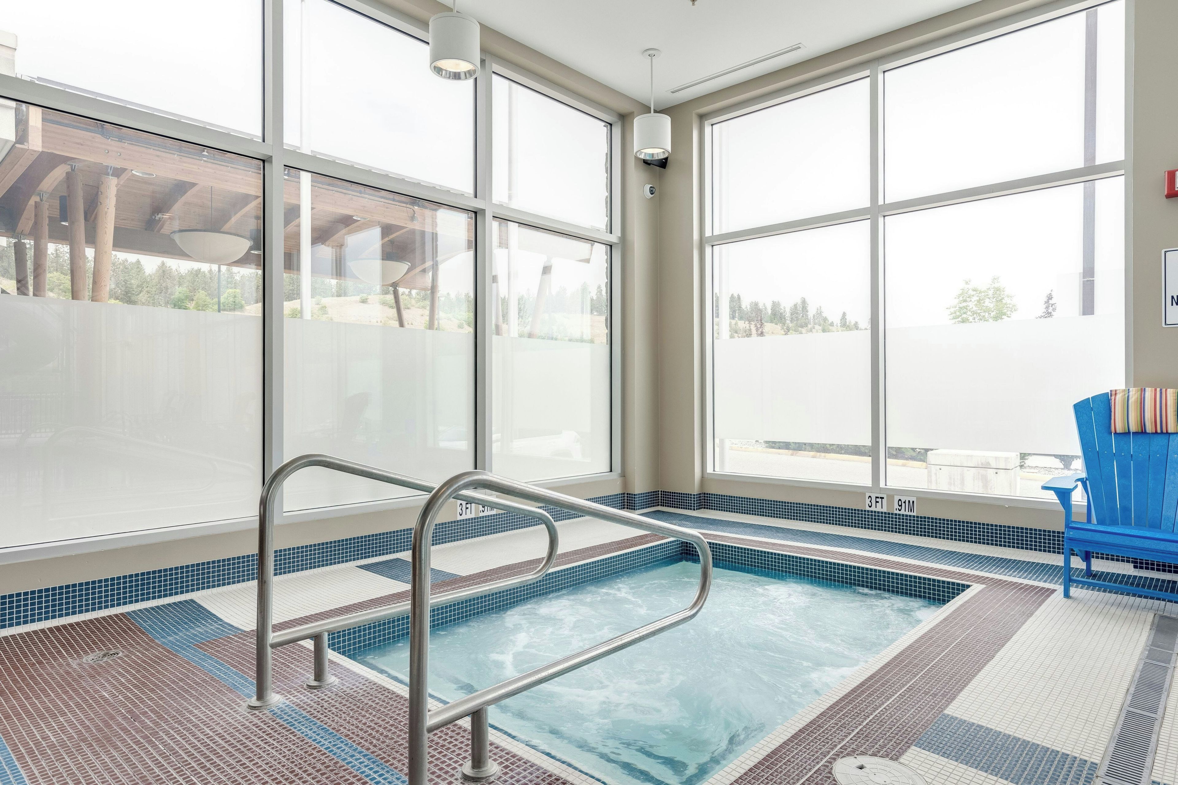 A swimming pool with a metal ladder, surrounded by large windows offering a view of the outdoors.