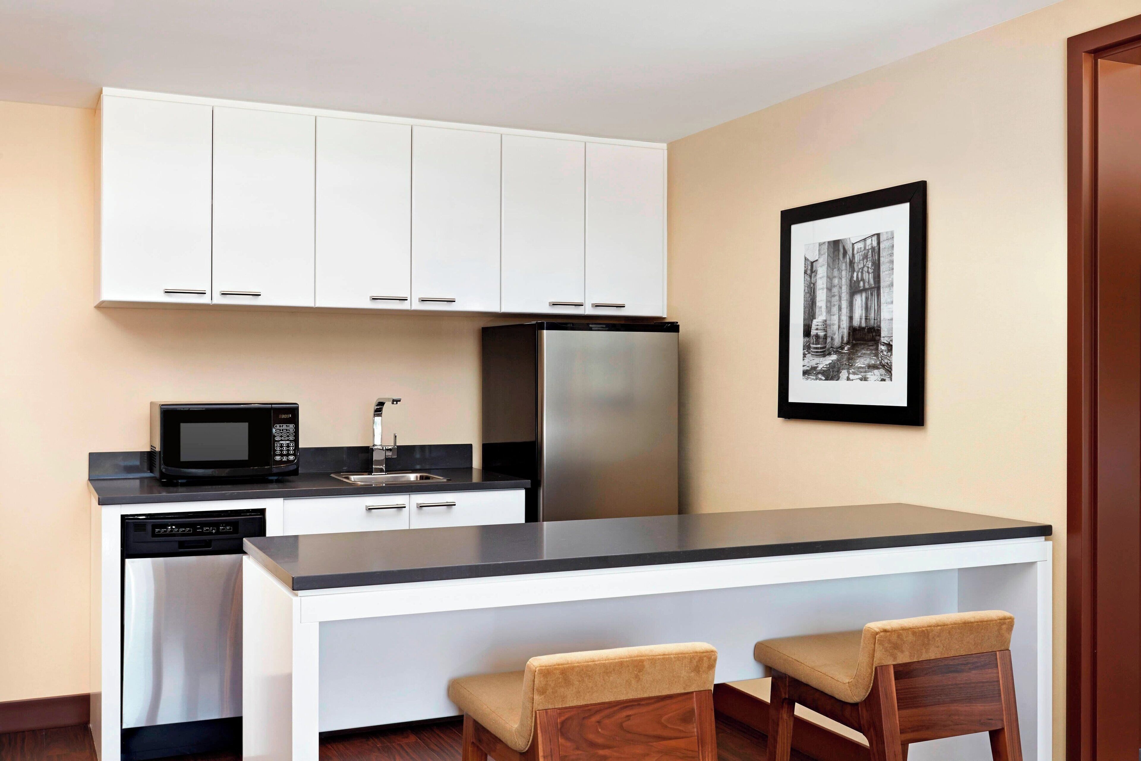 A compact kitchen with white cabinets, a stainless steel refrigerator, a microwave, and a dishwasher. There are two wooden bar stools at the counter.