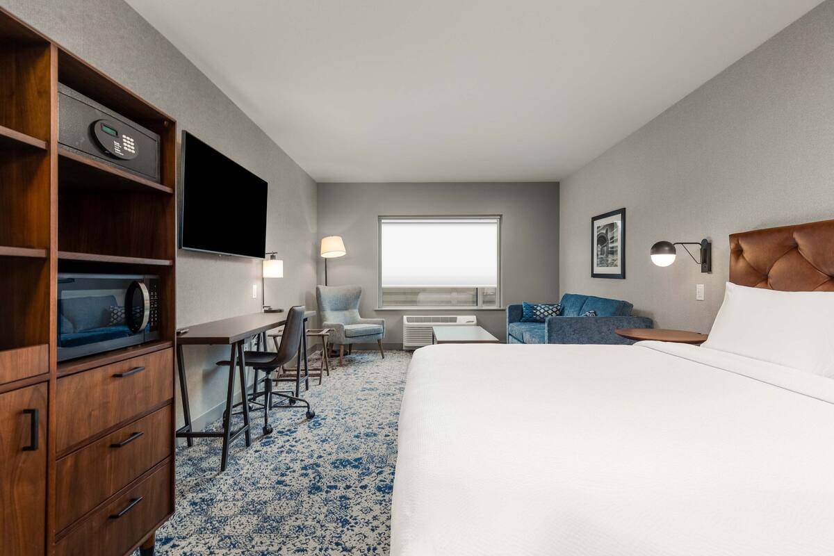 Quarto de hotel com cama, escrivaninha, cadeira, televisão, janela e luminária na parede.