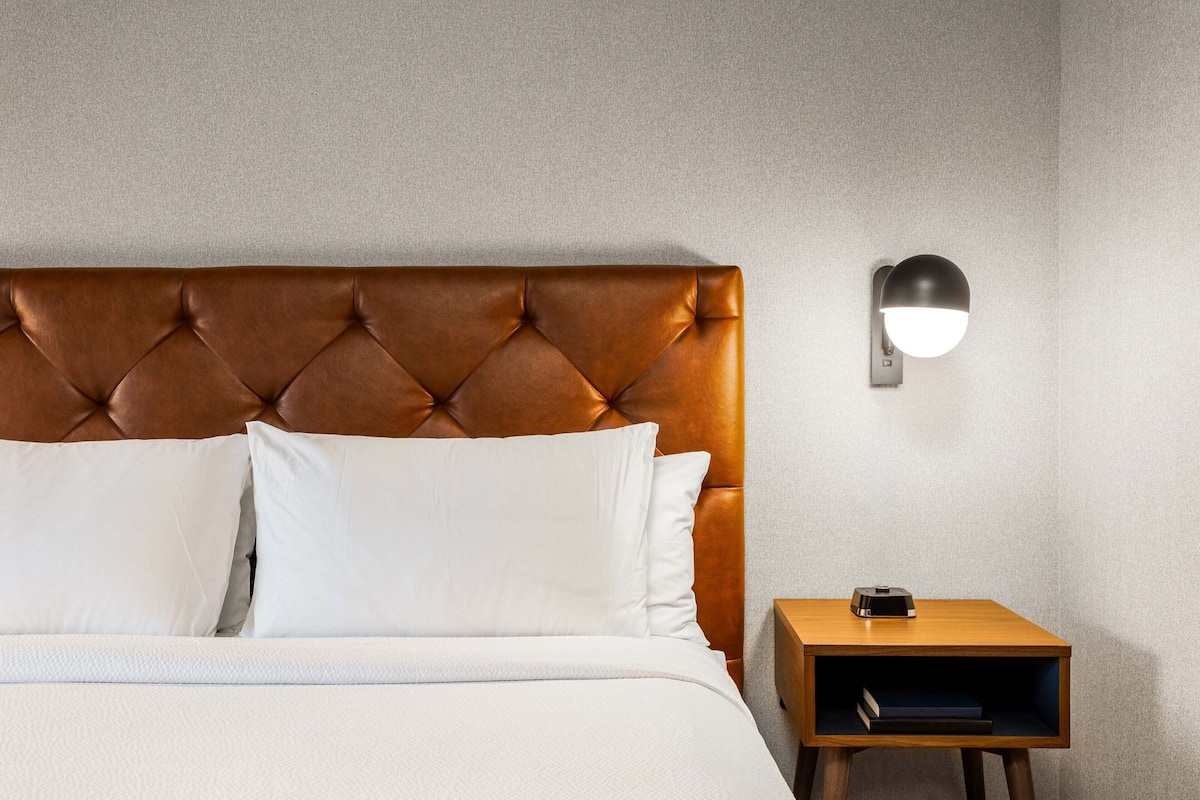 Cama bem arrumada com cabeceira estofada, mesa de cabeceira com luminária e pequena estante com livros.