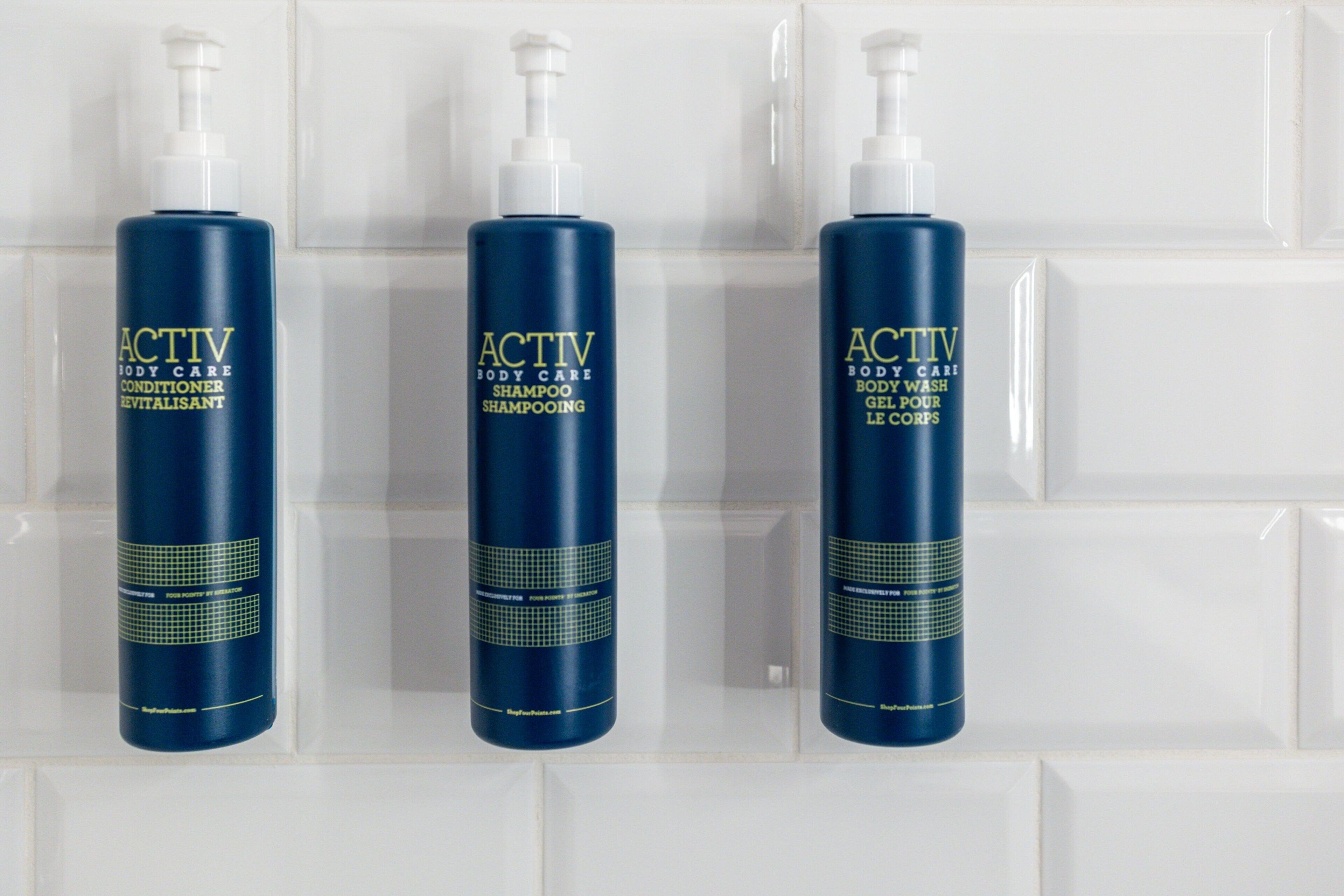Three bottles of Activ body care shampoo and conditioner on a white tiled wall.