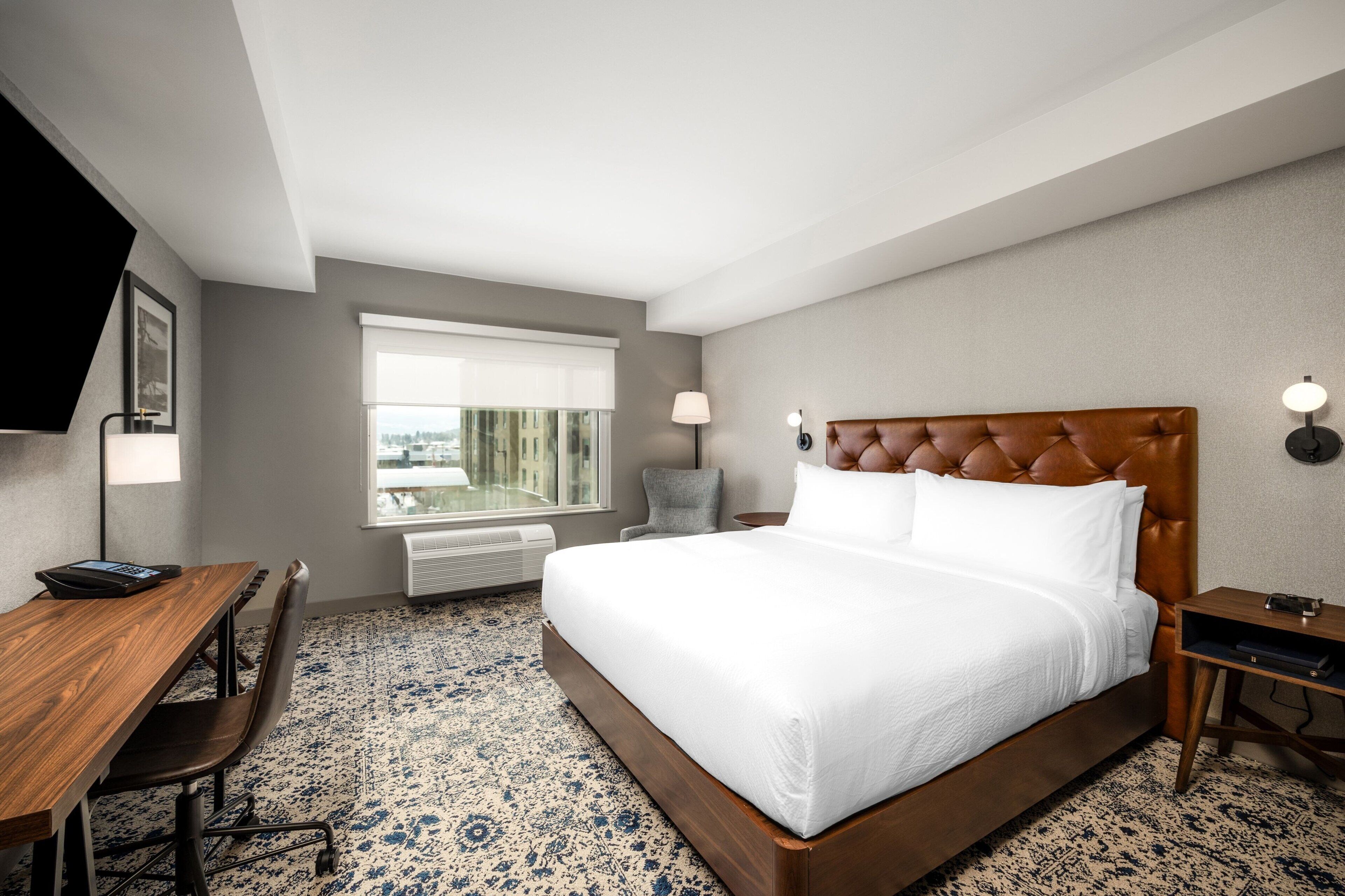 Habitación de hotel con una cama grande, un escritorio y una silla. Se ve una ventana con vistas a edificios y un televisor fijado a la pared.