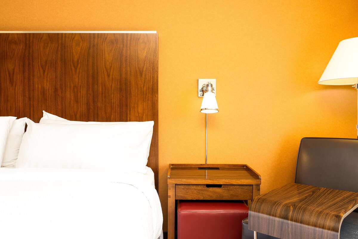 Habitación de hotel con cabecera de madera, mesita de noche con lámpara, un banco rojo y una mesita auxiliar de madera.