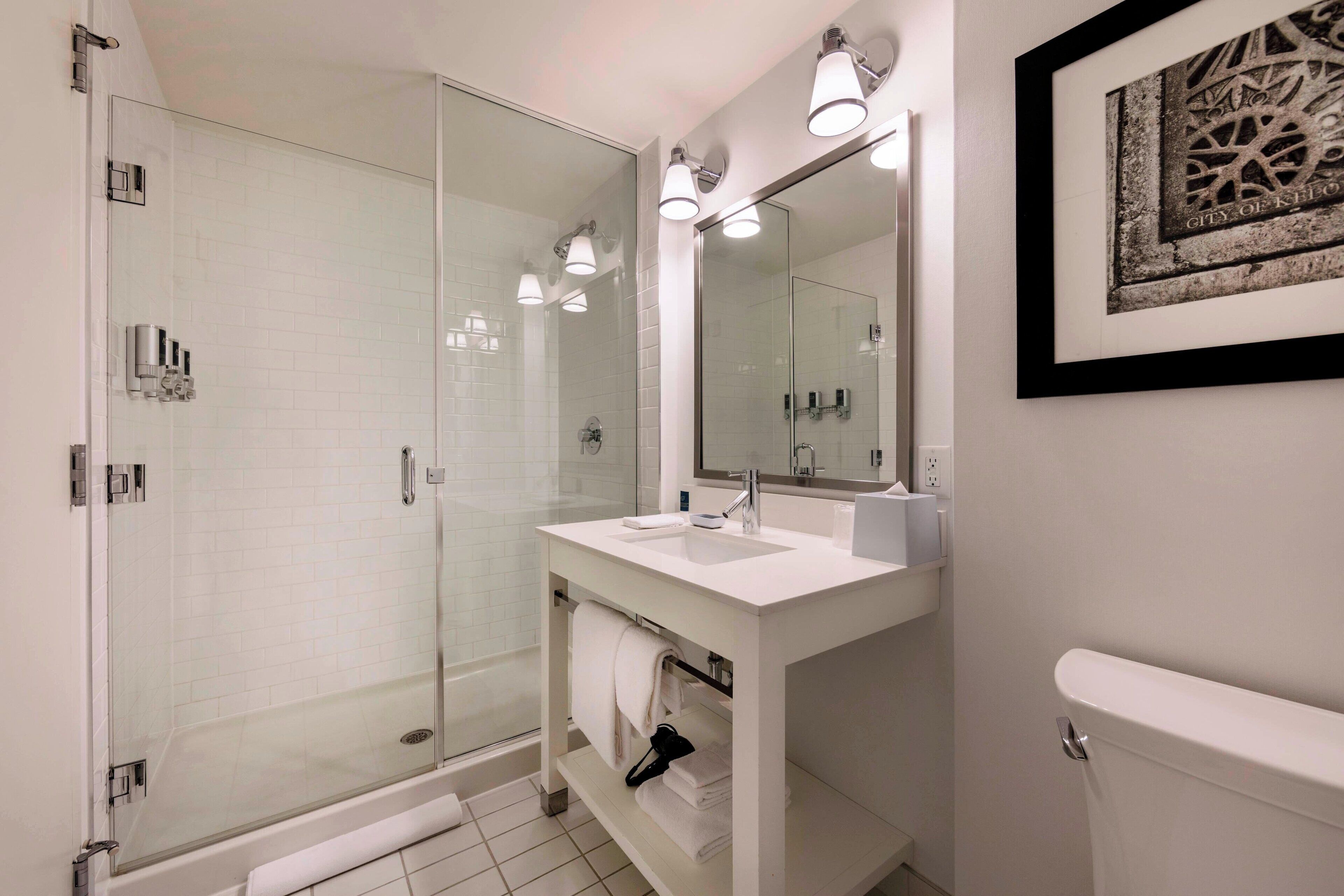 Une salle de bain moderne avec une douche à parois de verre, un lavabo blanc avec un miroir et une œuvre d’art encadrée au mur.