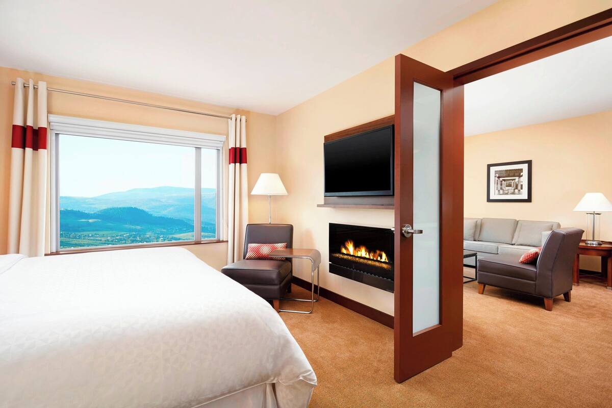 Quarto de hotel com cama, sofá, televisão, lareira e vista para as montanhas.