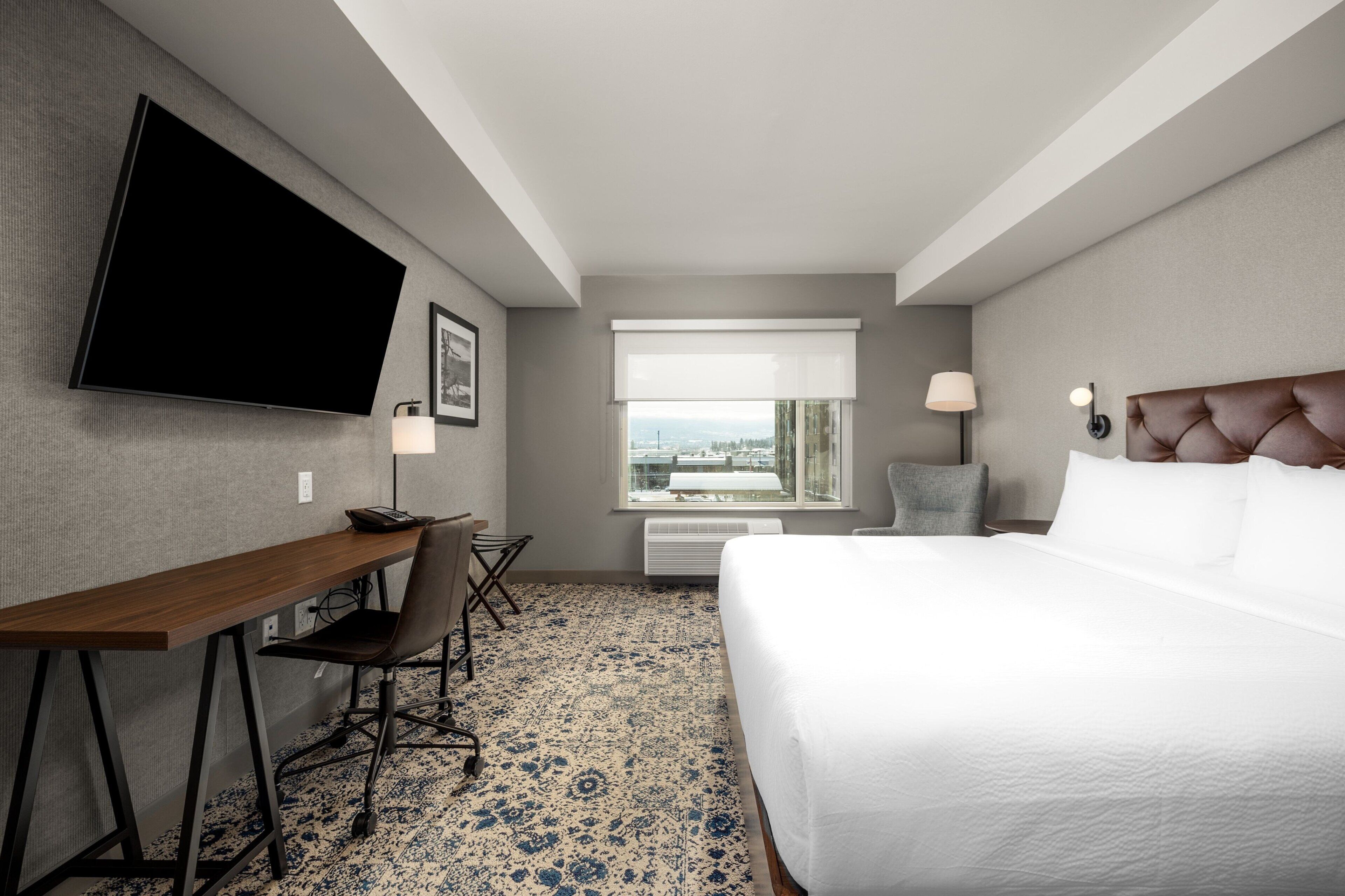 A hotel room with a bed, desk, chair, TV, window, and lamp.