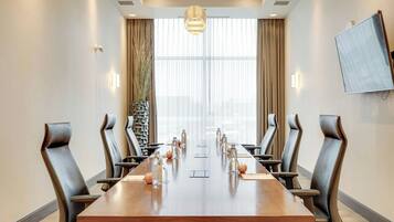 Ein Konferenzraum mit einem langen Tisch, Ledersesseln, einem an der Wand befestigten Fernseher und einem großen Fenster mit Vorhängen.