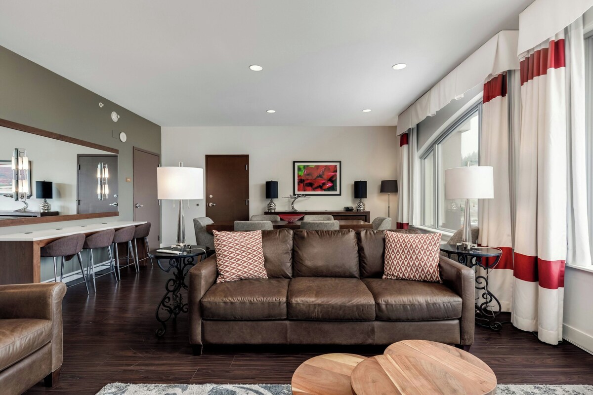 A modern living room with a brown leather sofa, a dining area with a bar, and a large window with curtains.