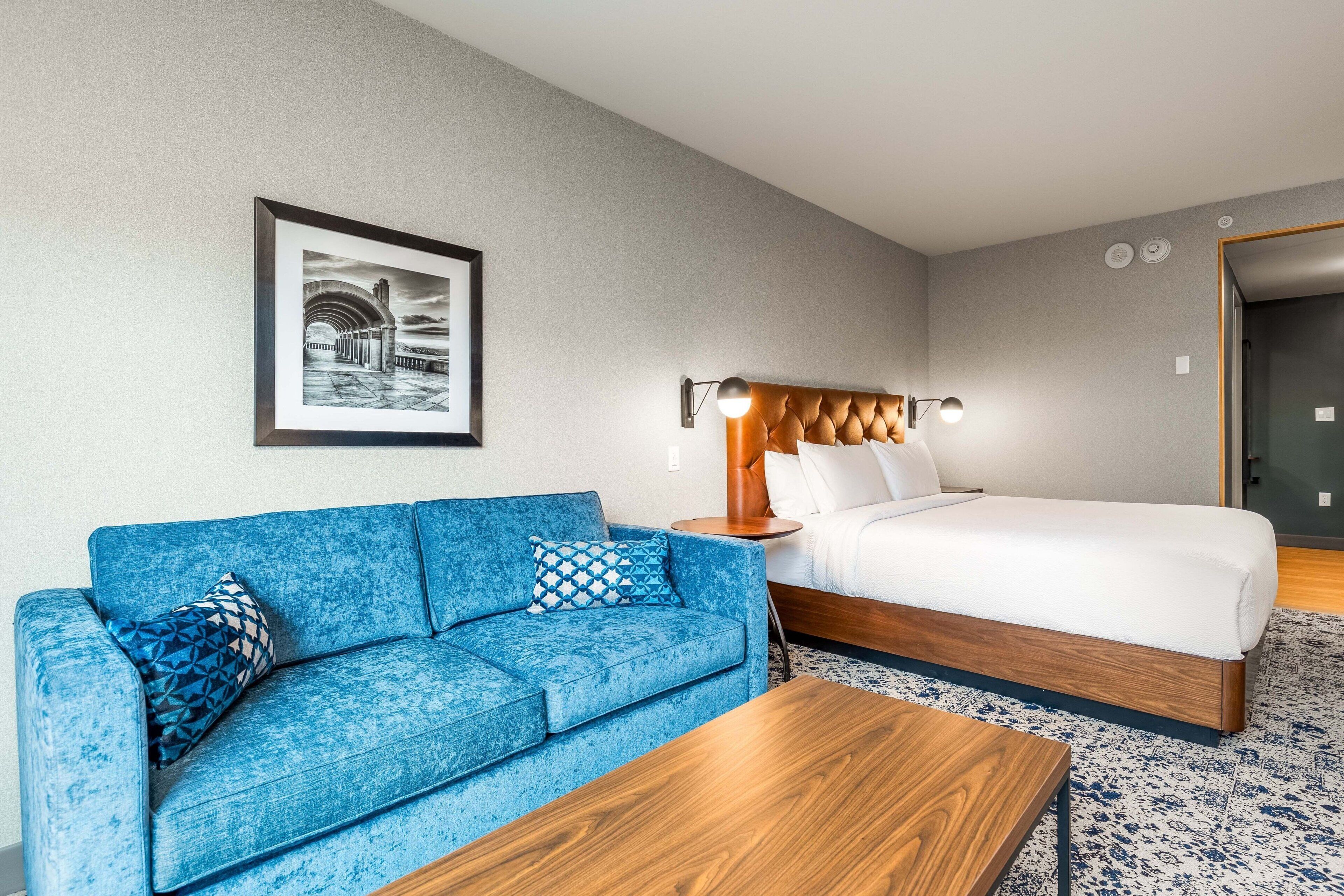 A modern hotel room with a blue sofa, a wooden coffee table, a bed with a wooden headboard, and a framed picture on the wall.