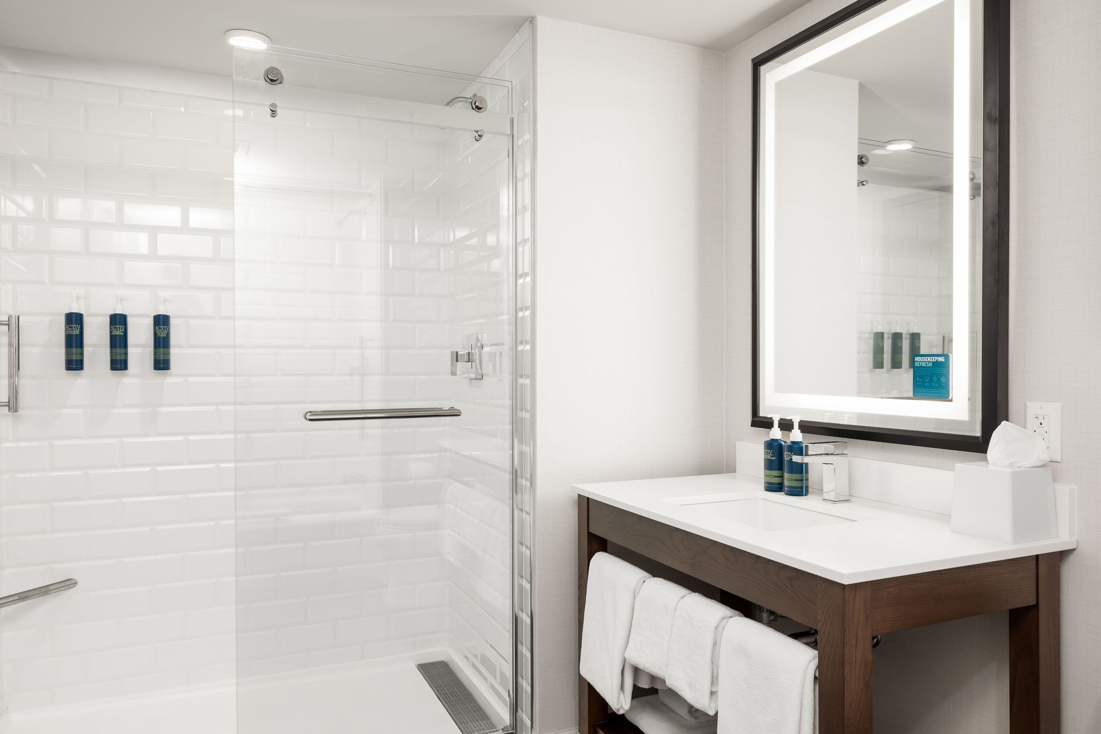 A modern bathroom with a glass shower enclosure, white tiled walls, a wooden vanity, and a large mirror.