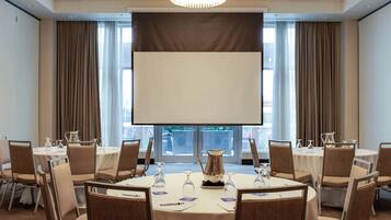 Een vergaderzaal met ronde tafels, stoelen, een projectiescherm en een groot raam met gordijnen.