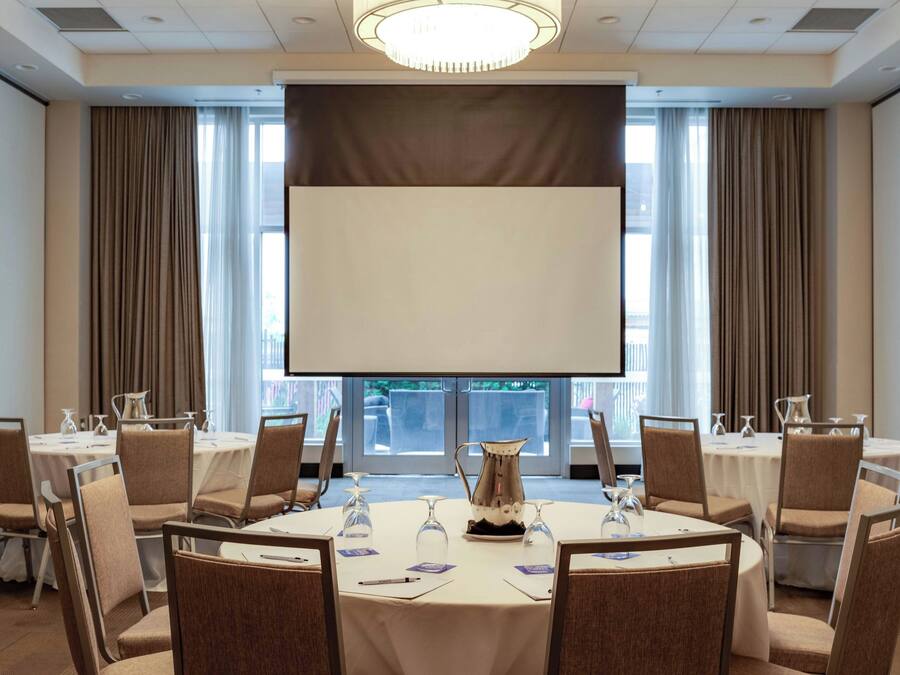 Een vergaderzaal met ronde tafels, stoelen, een projectiescherm en een groot raam met gordijnen.